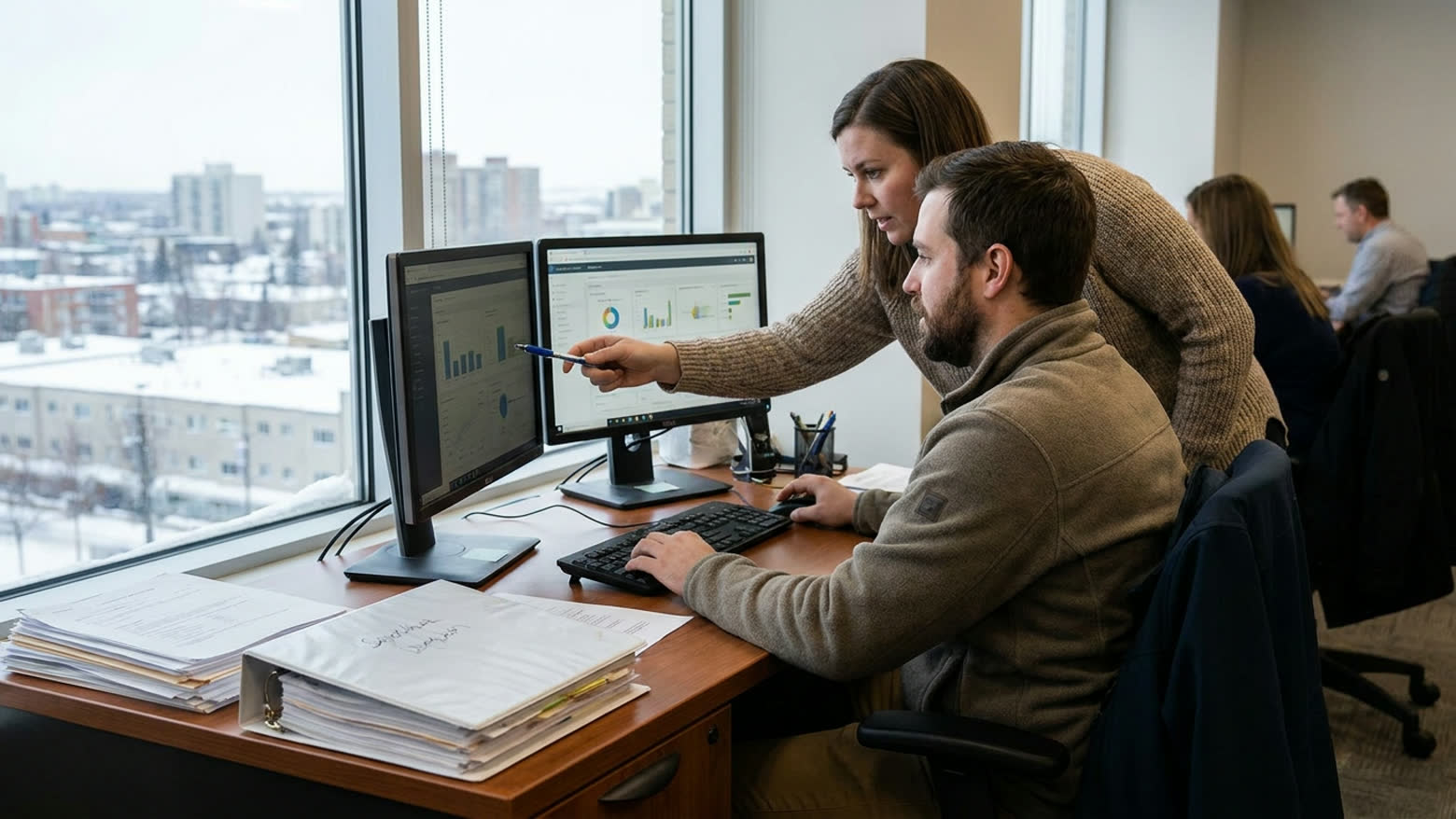 A planning analyst points at a chart on one of two monitors while a colleague follows, a snowy city through the window behind them.