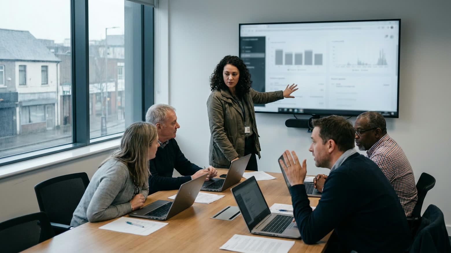 A woman presents a dashboard on a wall screen to four colleagues with laptops round a meeting table.